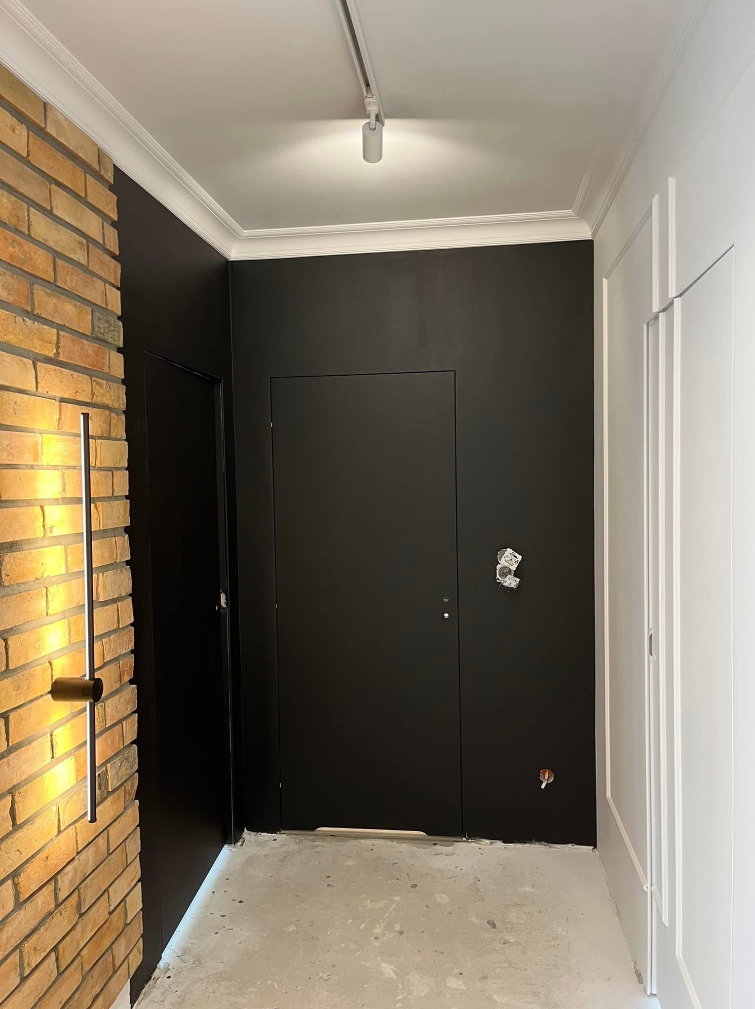 Nowoczesne wykończenie wnętrza: czarne ściany z oświetleniem LED przy ceglanej ścianie, białe listwy przysufitowe, minimalistyczne drzwi i surowe wykończenie podłogi.
