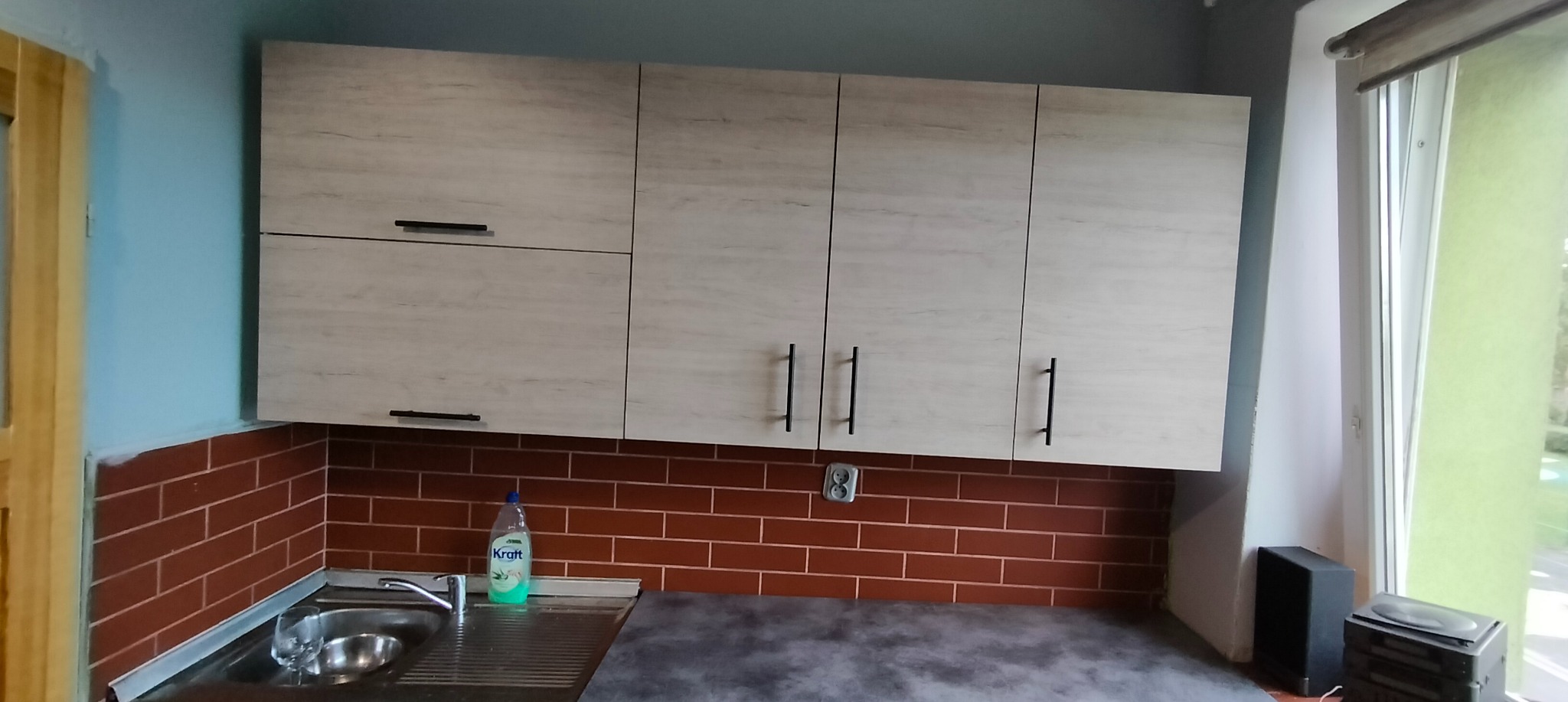 Kuchnia w Pieńsku po remoncie: jasne szafki wiszące z czarnymi uchwytami nad ceglaną ścianą, z widocznym zlewem i płynem do naczyń.