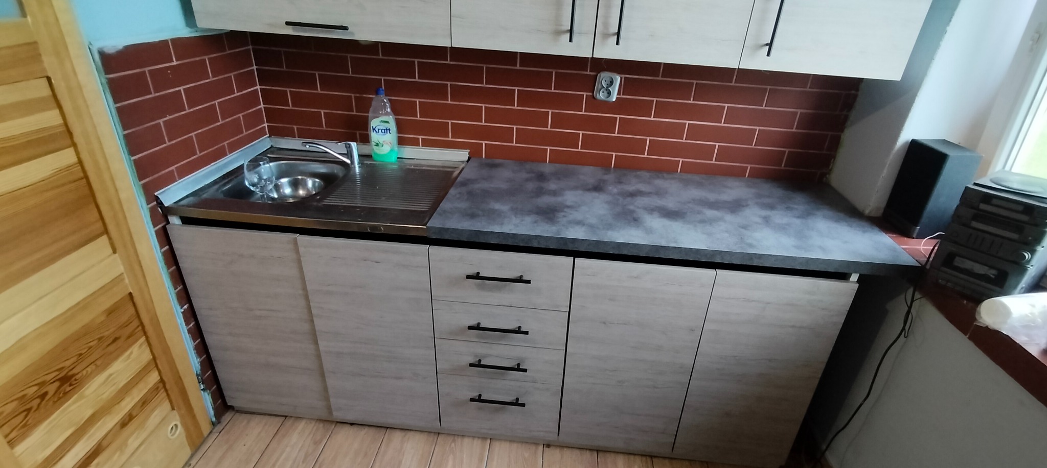 Fragment nowej zabudowy kuchennej z jasnymi frontami szafek, ciemnym blatem i ceglaną ścianą w tle, widoczny zlew ze szklanką i butelka płynu do naczyń.