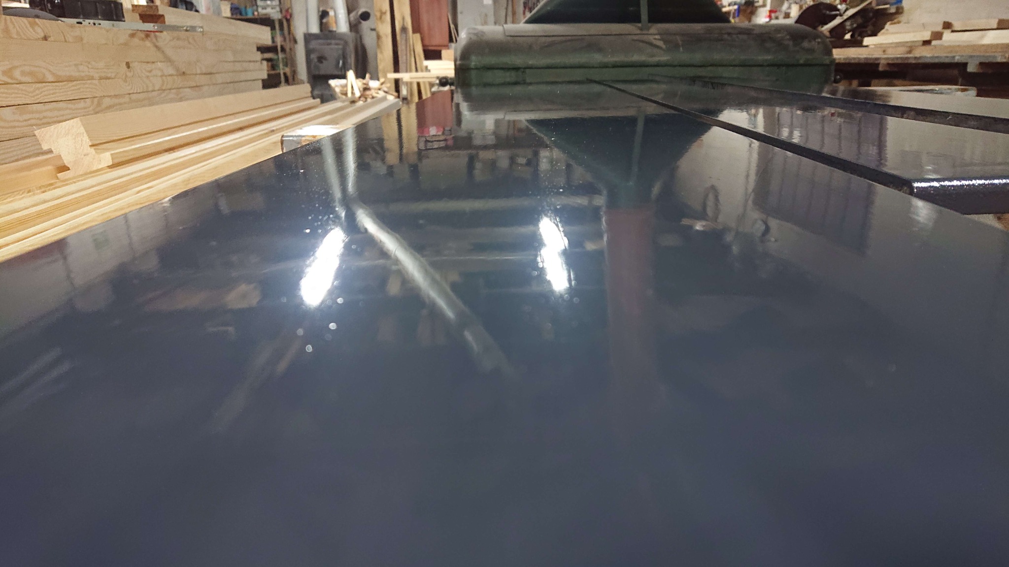 Błyszcząca, ciemnoszara powierzchnia, prawdopodobnie świeżo położony tynk, odbijająca światło lamp i elementy wyposażenia warsztatu, z widocznymi deskami w tle.