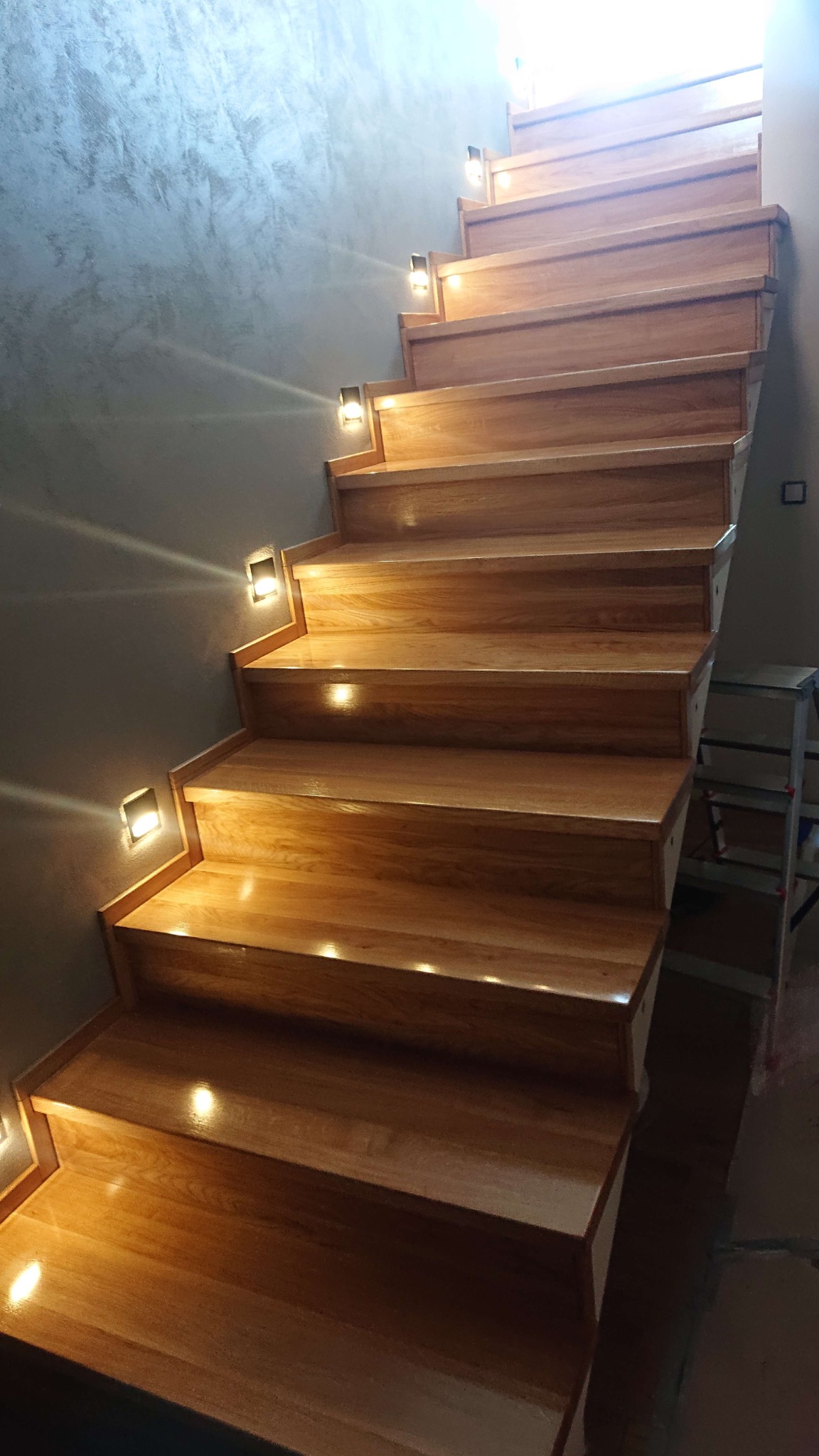Drewniane schody z oświetleniem punktowym w ścianie, widok z dołu, ściana wykończona dekoracyjnym tynkiem.