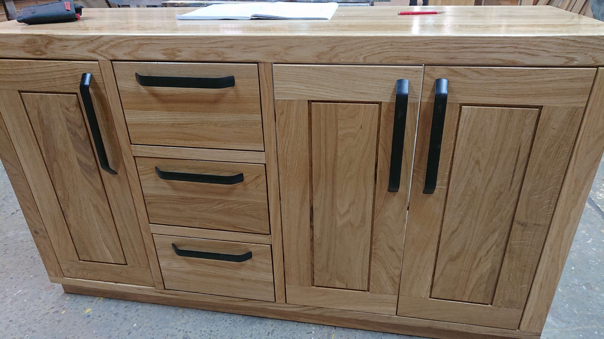 Drewniana szafka z trzema szufladami i dwojgiem drzwiczek, wyposażona w czarne, metalowe uchwyty, na blacie leżą notatnik i ołówek.