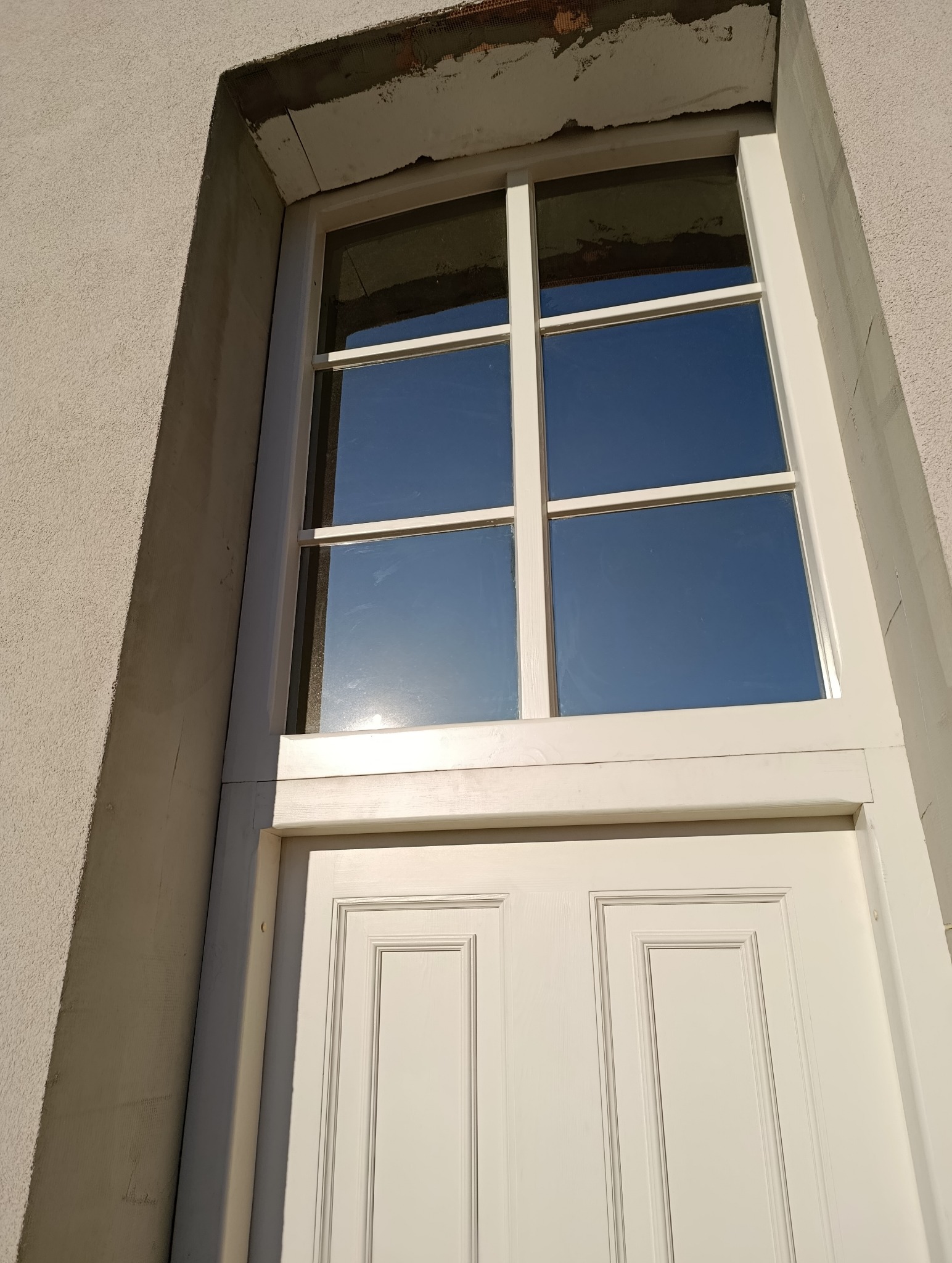 Białe, drewniane okno z szybami odbijającymi błękit nieba, osadzone w otynkowanej ścianie budynku. Widoczne ślady prac wykończeniowych nad oknem.