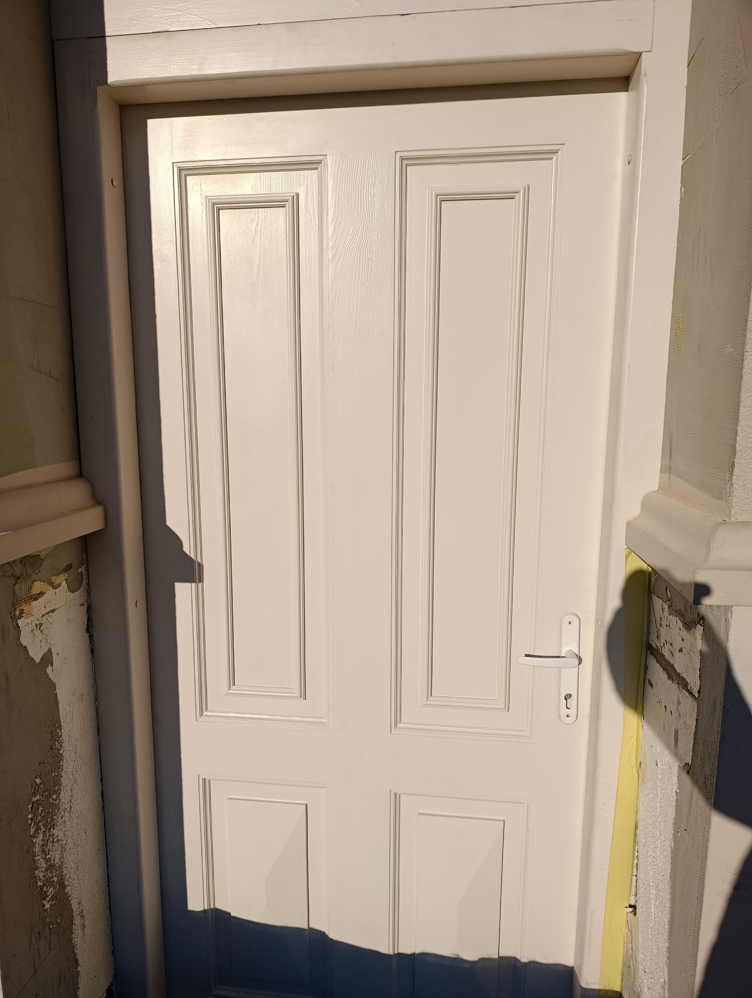 Białe, czteropanelowe drzwi wejściowe z klamką, osadzone w ościeżnicy. Widoczne fragmenty elewacji w trakcie prac wykończeniowych.