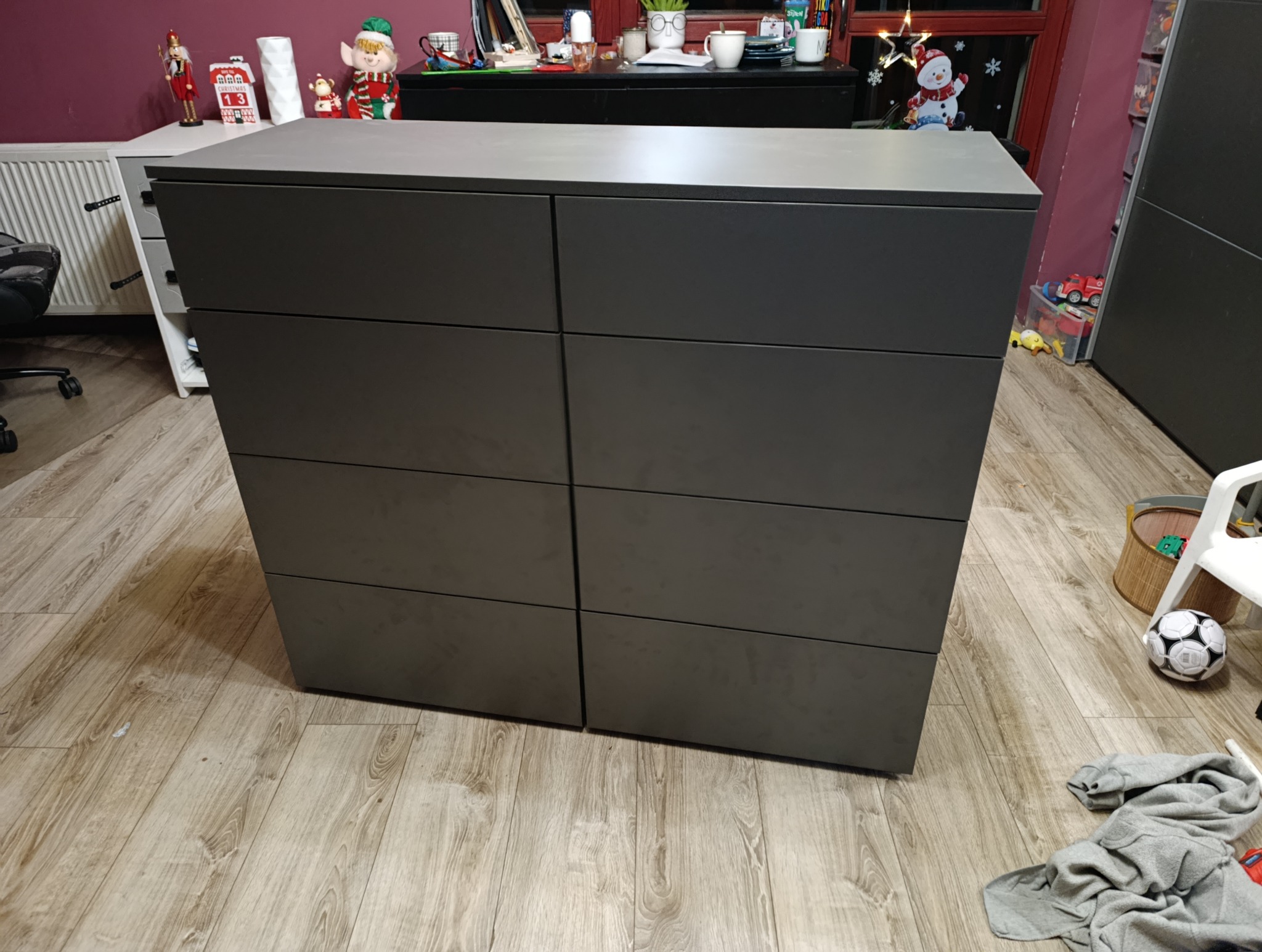 Szara komoda z sześcioma szufladami na drewnianej podłodze, z zabawkami w tle. Minimalistyczny design, proste linie, nowoczesny wygląd.