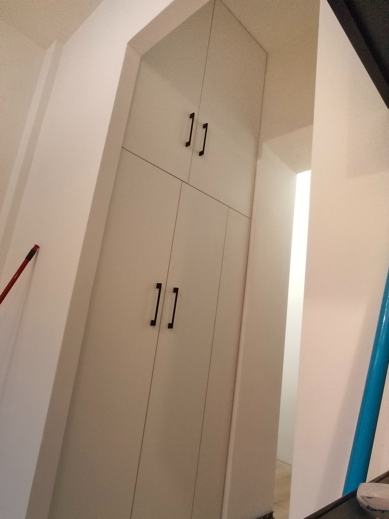 Wysoka, biała szafa na wymiar z czarnymi uchwytami, wkomponowana w narożnik pomieszczenia. Widoczny fragment ściany i przejście do innego pokoju.