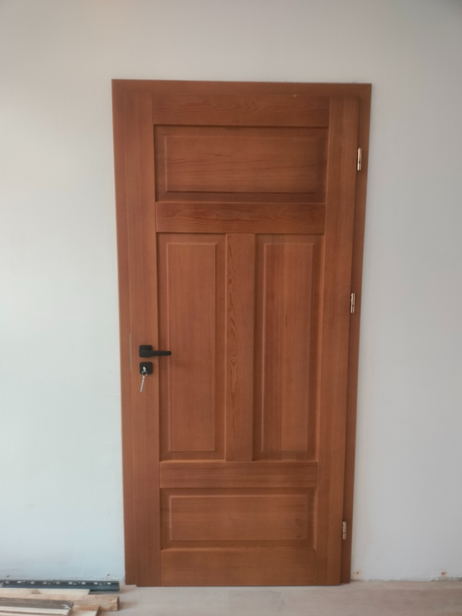 Zamknięte, drewniane drzwi wewnętrzne w odcieniach brązu z czarną klamką i kluczem w zamku, na tle białej ściany. Widoczne listwy przypodłogowe.