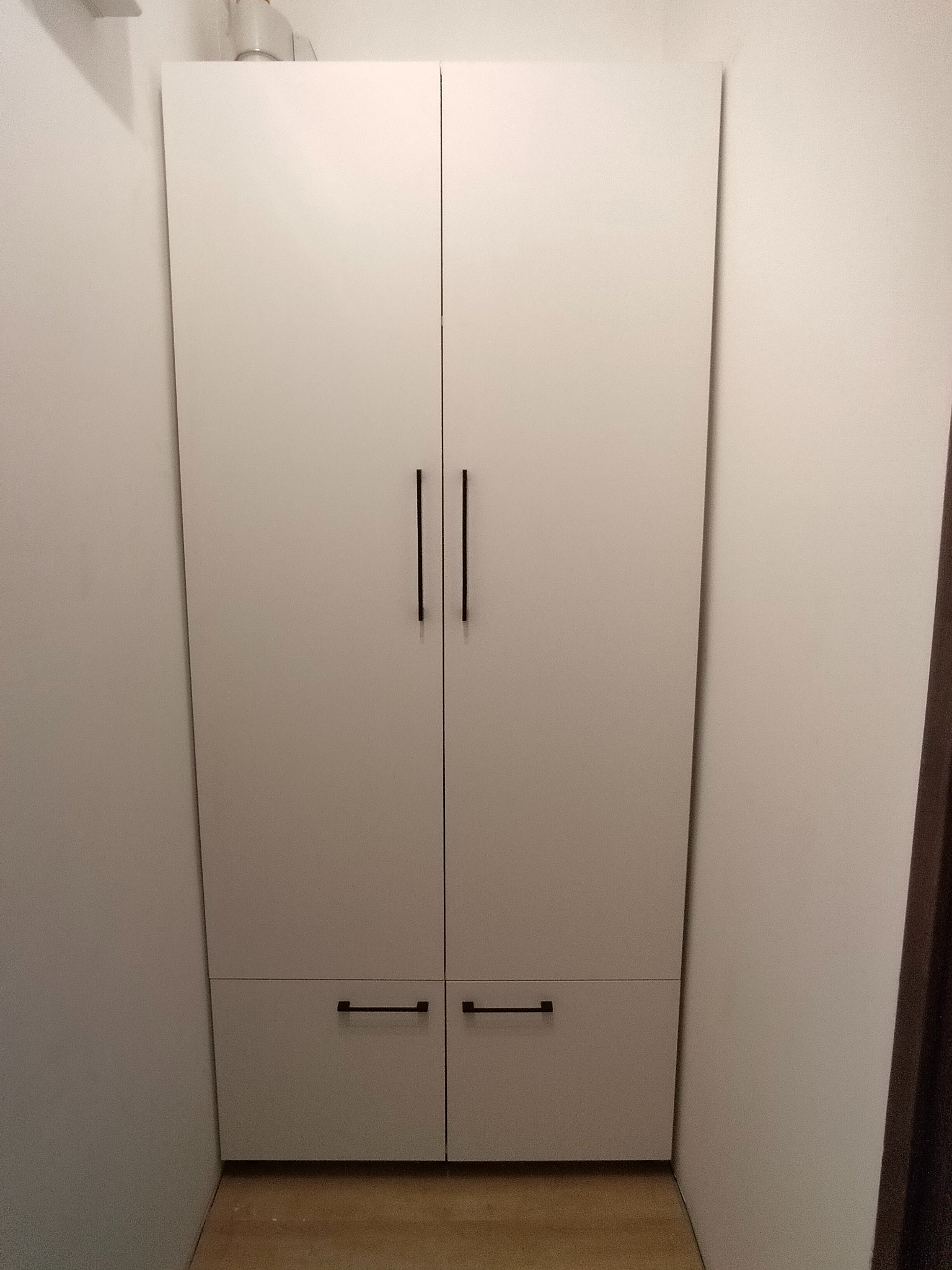 Biała, dwudrzwiowa szafa z ciemnymi, minimalistycznymi uchwytami, wpasowana w narożnik pokoju. Prosty design, jasne wnętrze, widoczna podłoga.