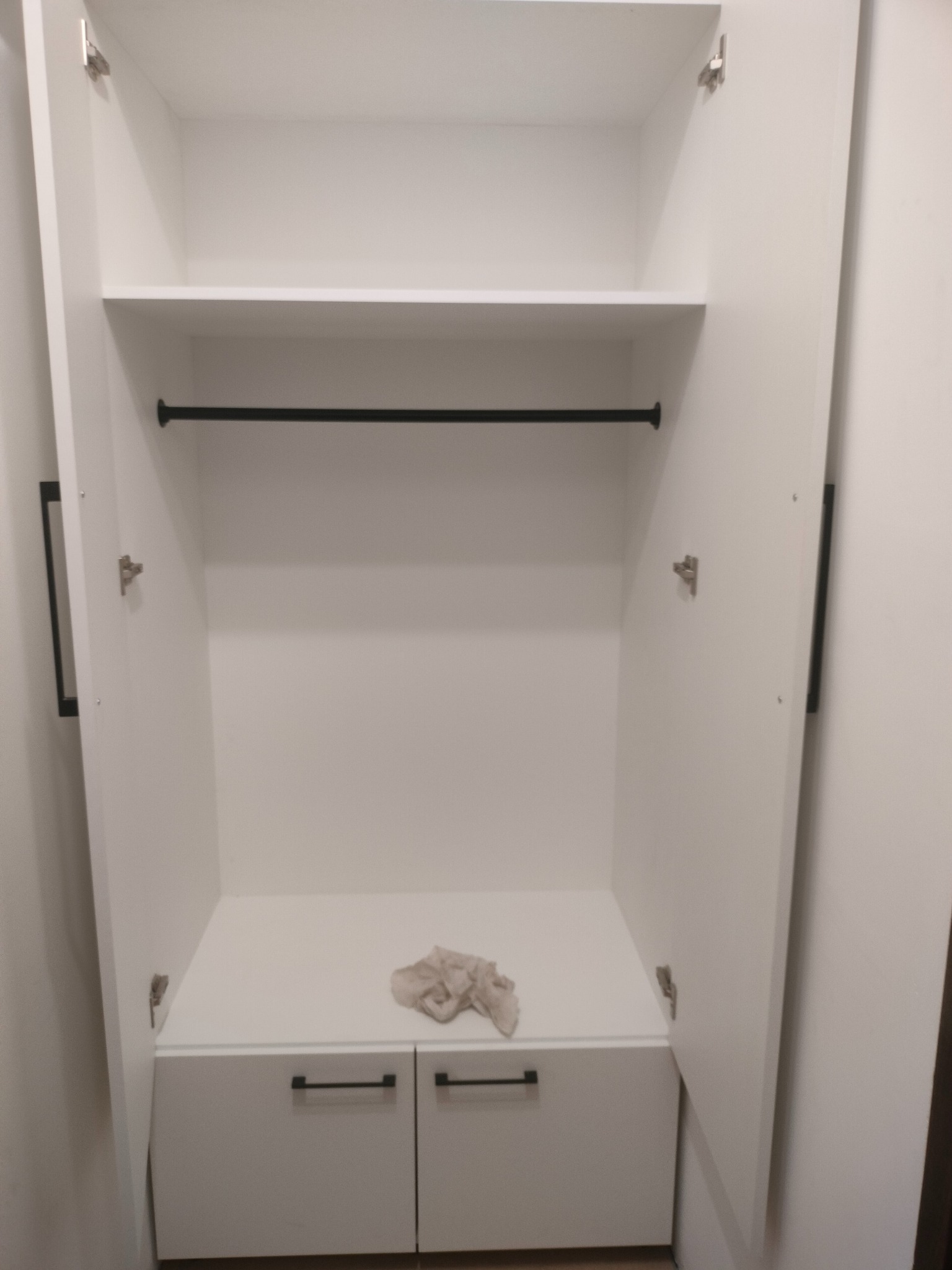 Wnętrze białej szafy na wymiar z otwartymi drzwiami, widoczna półka, drążek na ubrania i dwie szuflady z czarnymi uchwytami. Na dolnej półce leży szmatka.