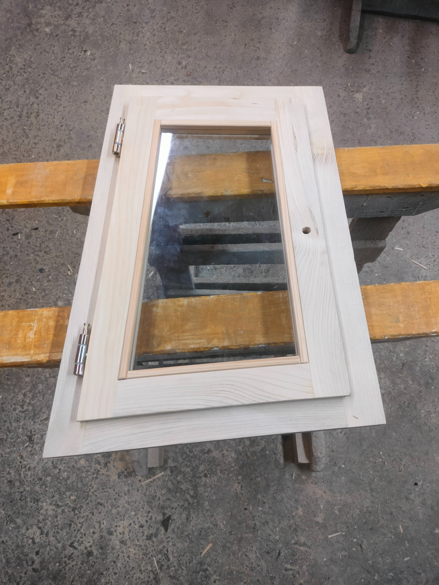 Drewniane, surowe skrzydło szafki z szybą i zawiasami, leżące na drewnianym stelażu. Widoczne usłojenie drewna i metalowe okucia. Element meblowy w warsztacie.