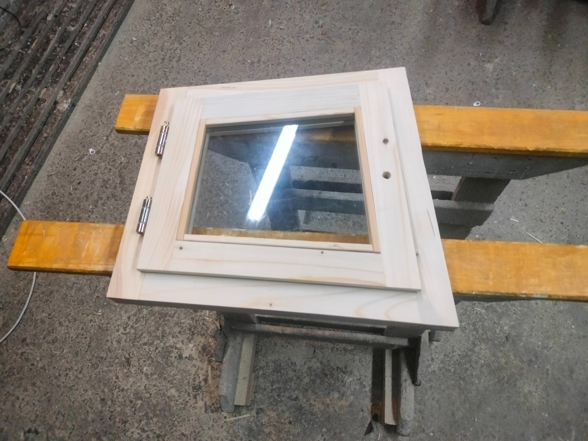 Surowa, drewniana rama okienna z zawiasami, leżąca na żółtych deskach. Widoczne otwory na śruby i odbicie światła w szybie. Szary beton w tle.