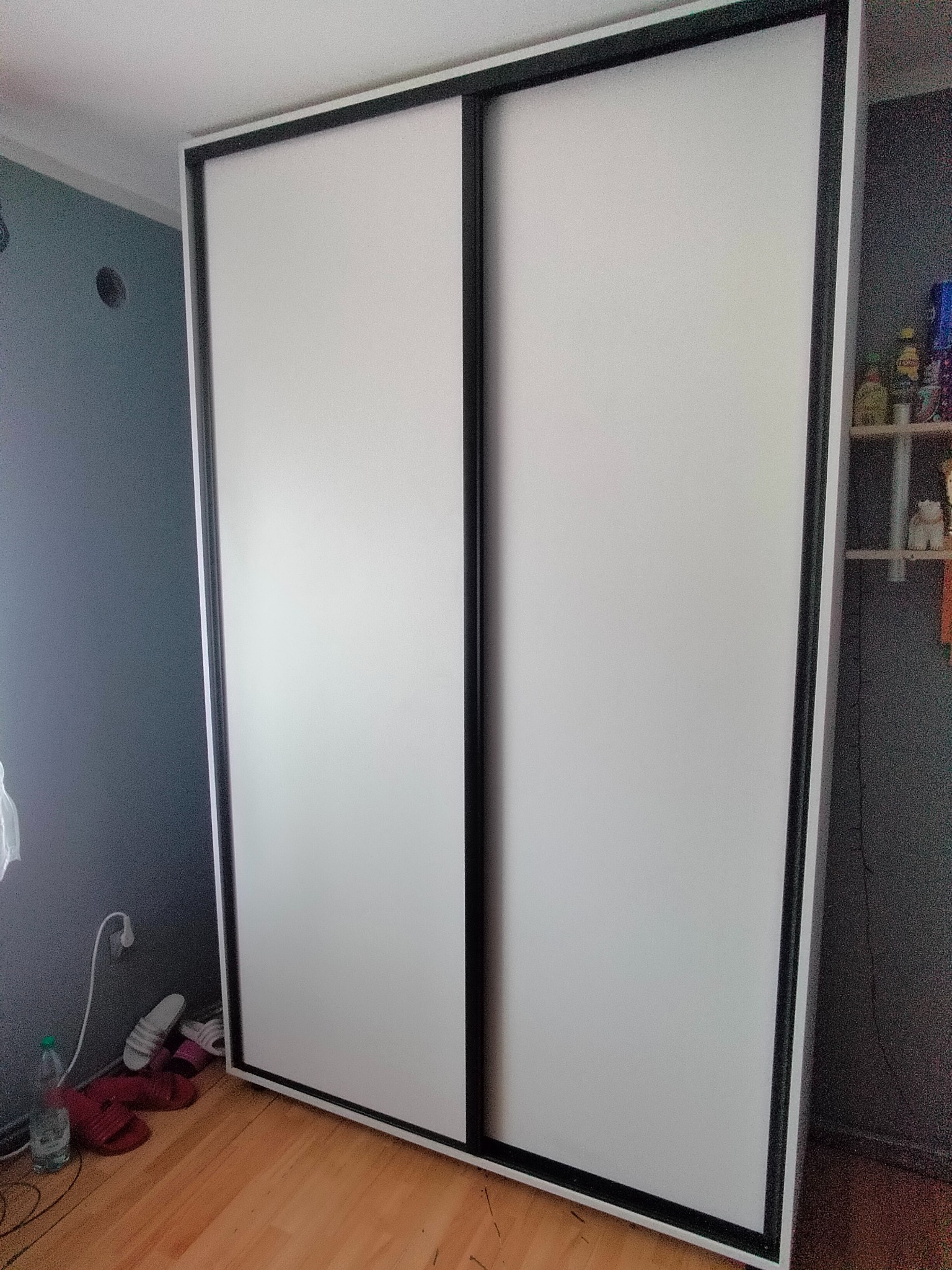 Nowoczesna szafa przesuwna z białymi frontami i czarnymi obramowaniami, stojąca na drewnianej podłodze w pokoju o szarych ścianach. Widoczne detale: gniazdko, półka.