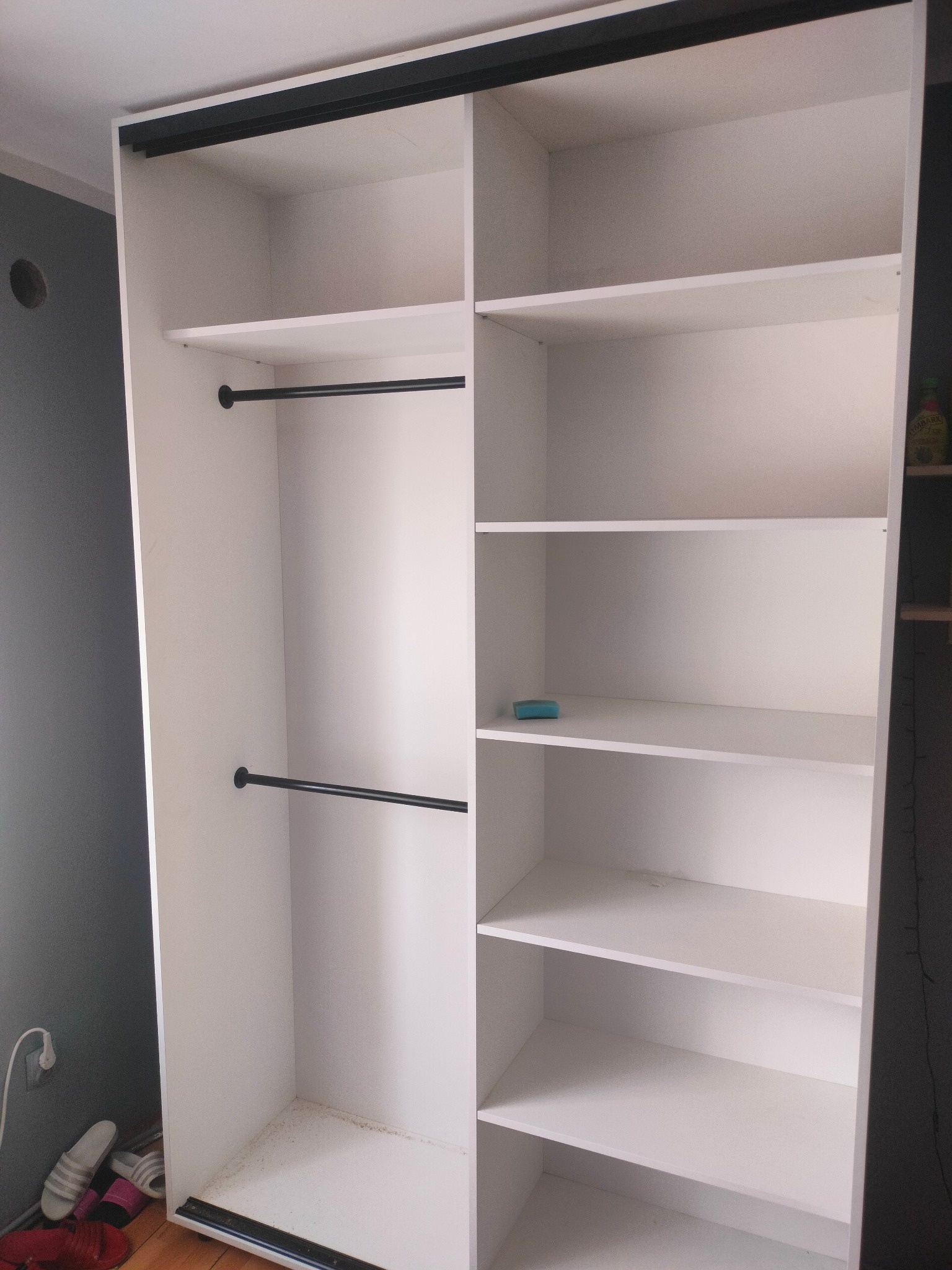 Biała szafa na wymiar z czarnymi detalami, widoczne półki i drążki na ubrania. Minimalistyczny design, wnętrze szafy puste, gotowe do użytku. Ujęcie z bliska.