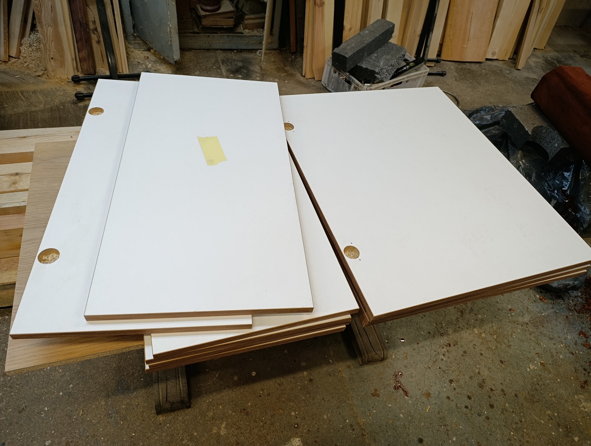 Białe, lakierowane fronty meblowe z nawierconymi otworami na zawiasy, ułożone warstwami w warsztacie. Na jednym z frontów widoczna żółta taśma maskująca.