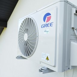 El-Rem Sp. z o. o. - Biała, zewnętrzna jednostka klimatyzacji marki Gree na ścianie budynku z widocznym logo i elementami instalacji. Widok z dołu i boku.
