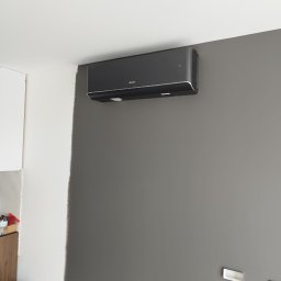 El-Rem Sp. z o. o. - Czarny klimatyzator Gree zamontowany na szarej ścianie w pomieszczeniu z elementami białej ściany i widocznym fragmentem mebli. Widoczne gniazdka elektryczne.
