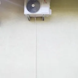 Biała jednostka zewnętrzna klimatyzacji Rotenso zamontowana na jasnej elewacji budynku z widoczną instalacją. Ujęcie z dołu, fragment dachu i desek podłogowych.