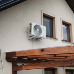 Biały klimatyzator Rotenso zamontowany na elewacji domu z beżowym tynkiem, widoczna aluminiowa drabina i drewniana pergola.