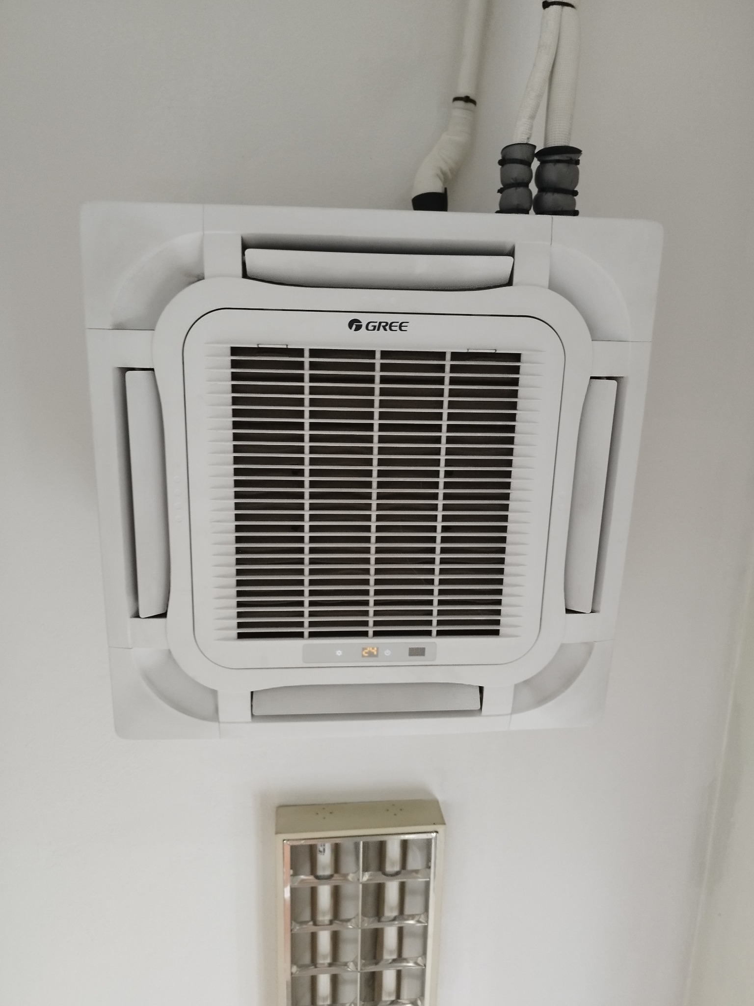 Sufitowa klimatyzacja kasetonowa marki Gree, widok z dołu, na białym suficie. Widoczne rury instalacyjne i lampa sufitowa. Ustawiona temperatura 24 stopnie.