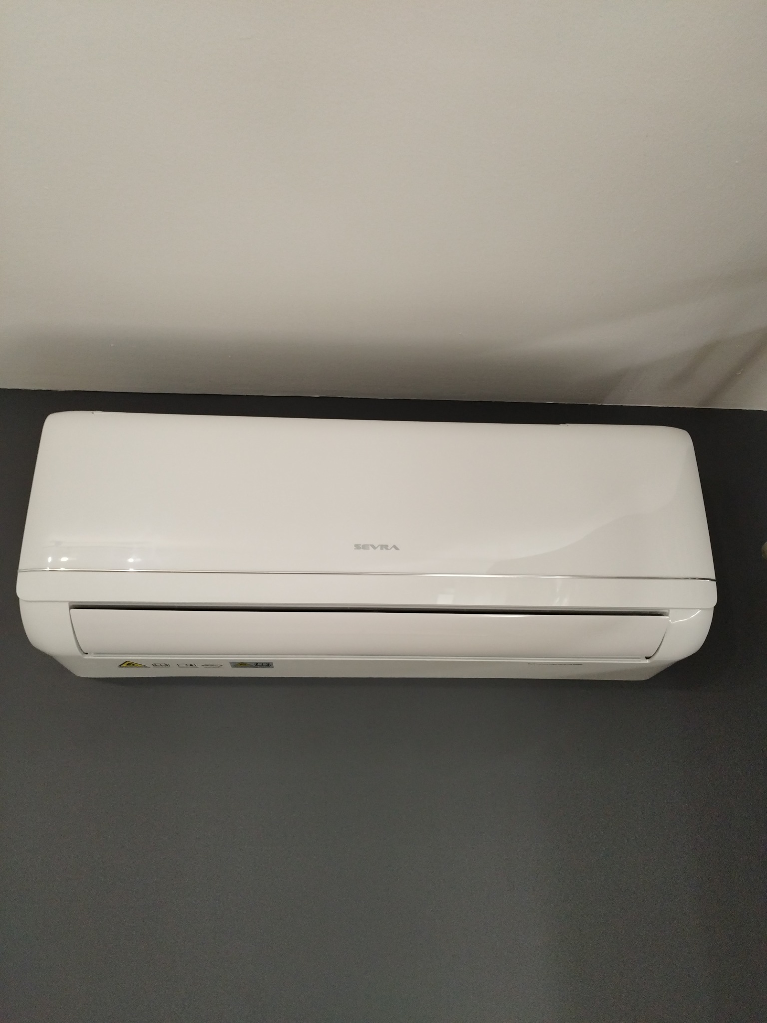 Biała klimatyzacja ścienna marki Sevra, zamontowana na szarej ścianie, widok z dołu, oświetlenie naturalne.