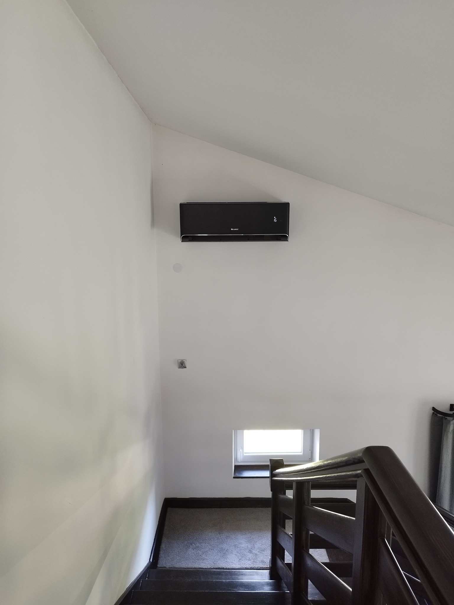 Czarna klimatyzacja Gree zamontowana na białej ścianie poddasza, widoczna z dołu schodów. Małe okno w tle i fragment drewnianej balustrady.