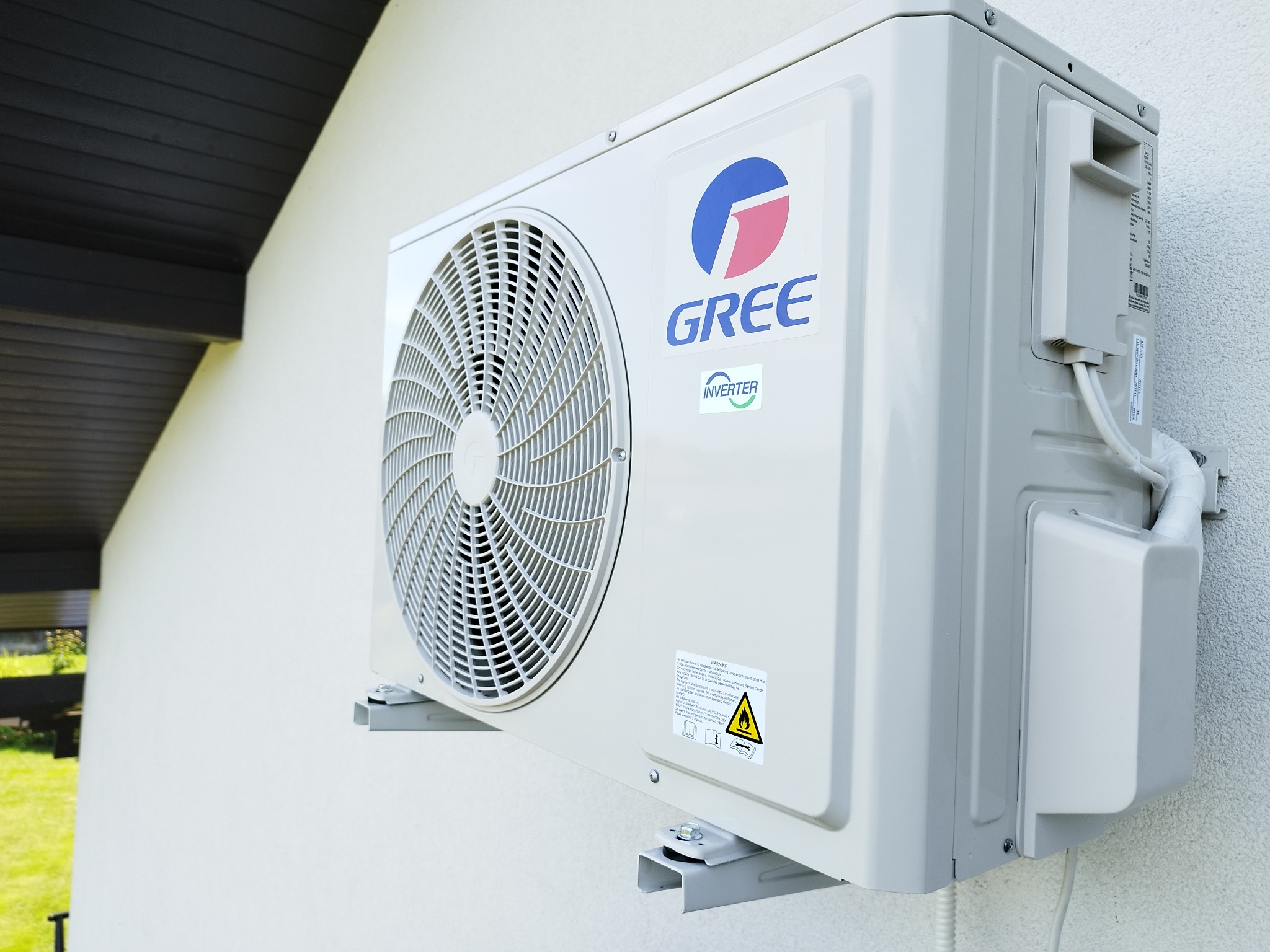 Biała, zewnętrzna jednostka klimatyzacji marki Gree na ścianie budynku z widocznym logo i elementami instalacji. Widok z dołu i boku.