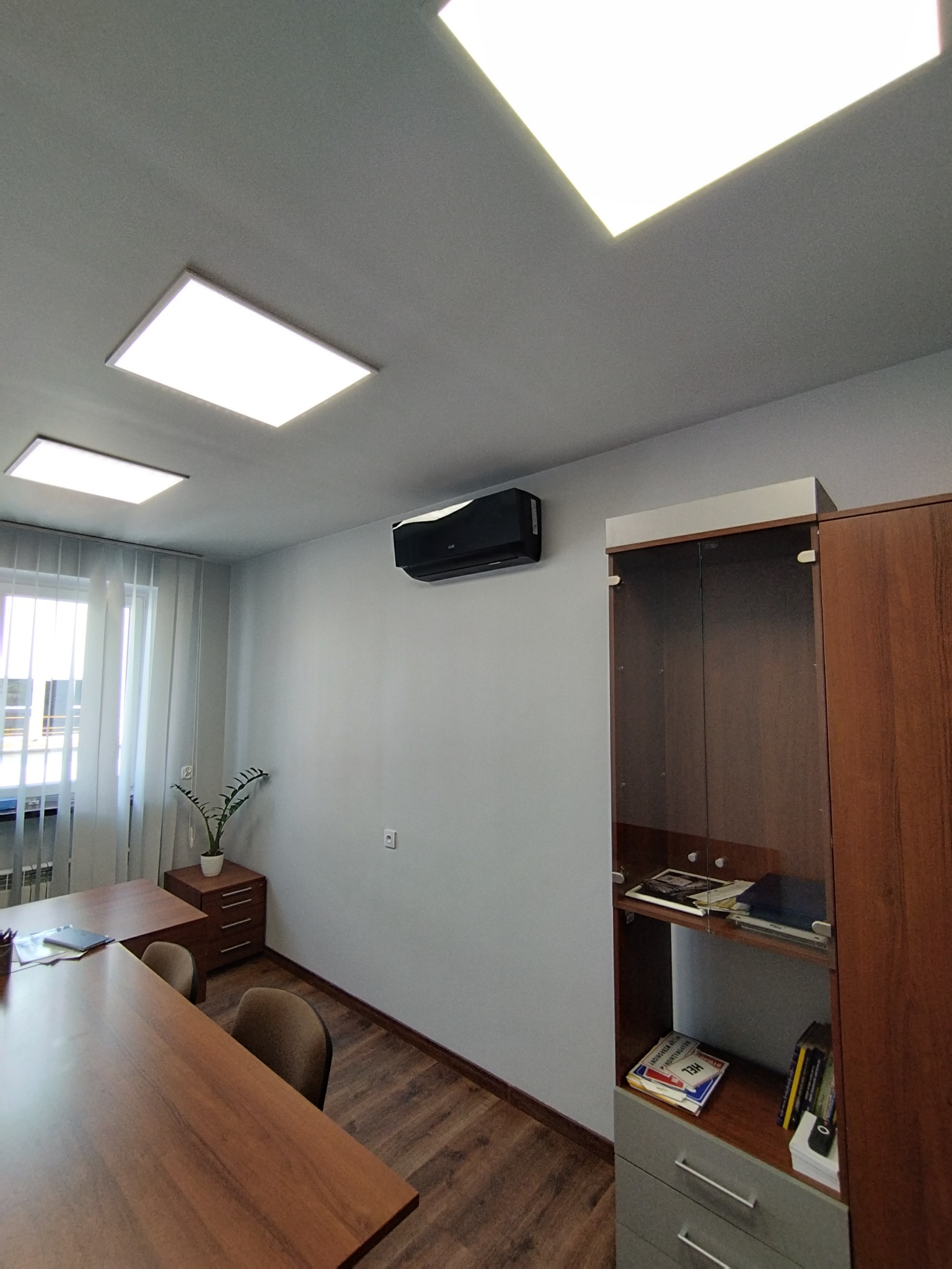 Nowoczesne biuro z czarną klimatyzacją ścienną, oświetleniem LED i drewnianymi meblami. Widok na okno z żaluzjami i roślinę doniczkową.