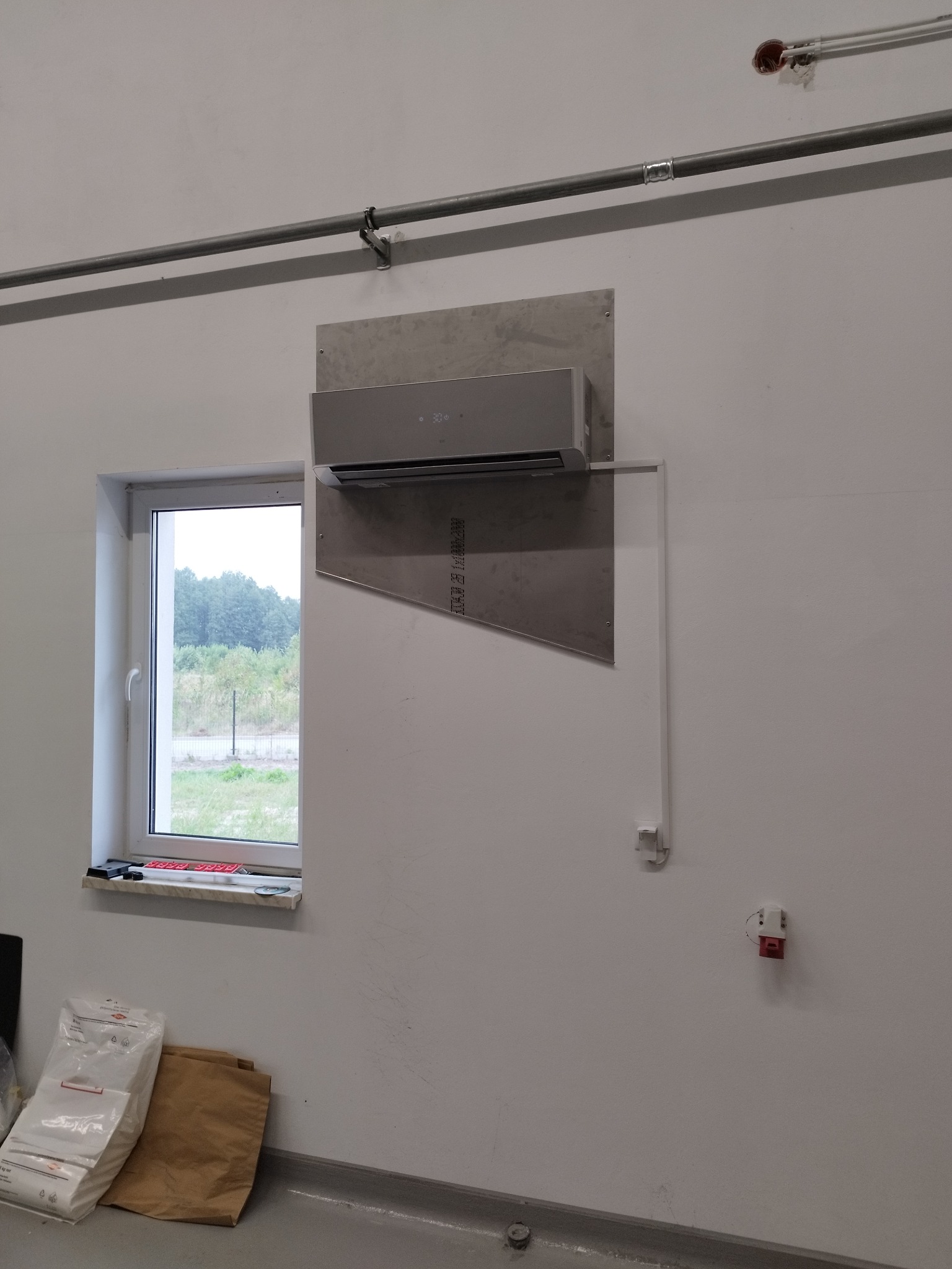 Szary klimatyzator zamontowany na metalowej płycie na białej ścianie w pomieszczeniu z oknem i instalacjami elektrycznymi. Widoczne worki na podłodze.
