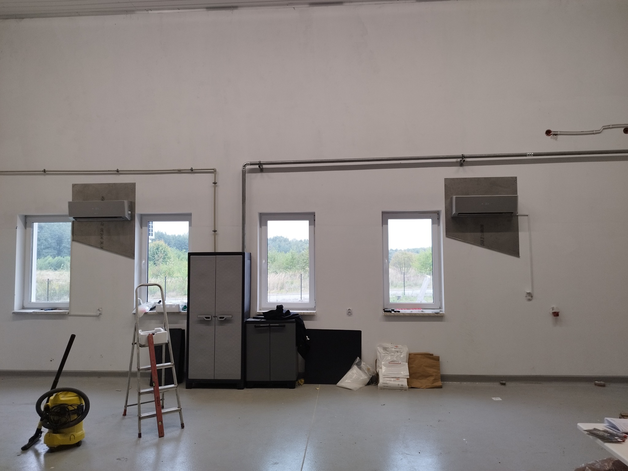 Montaż klimatyzacji w nowym, przestronnym pomieszczeniu. Widoczne dwa klimatyzatory, drabina, odkurzacz przemysłowy i materiały budowlane.