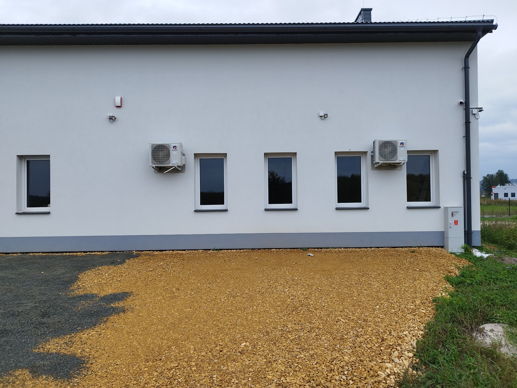Biała fasada budynku z dwoma klimatyzatorami Gree zamontowanymi między oknami. Widoczne instalacje elektryczne i fragment terenu z kamieniami.