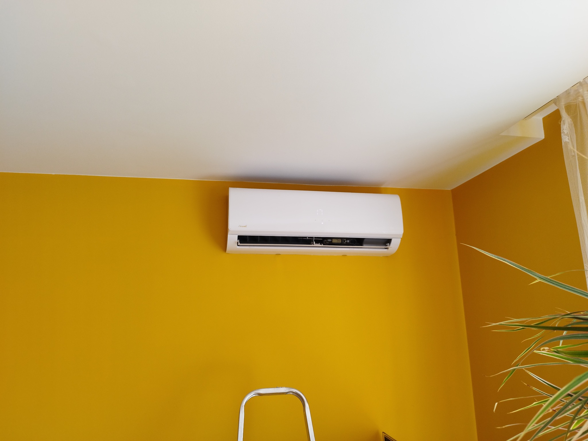 Biała klimatyzacja na żółtej ścianie w pokoju, widoczna drabina i roślina doniczkowa. Ujęcie z dołu, z fragmentem sufitu.