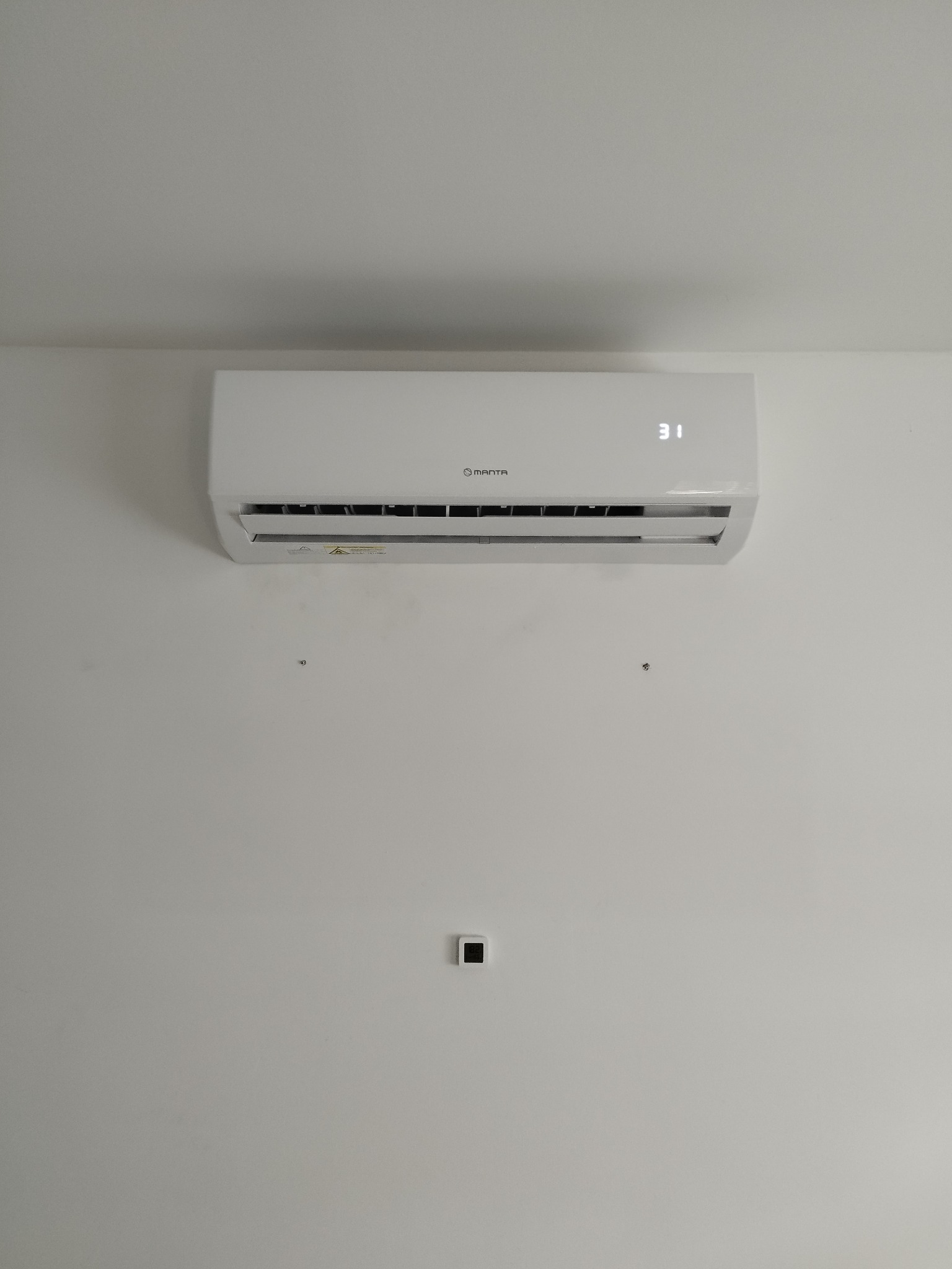 Biała klimatyzacja Manta z wyświetlaczem 31 stopni Celsjusza zamontowana na białej ścianie. Widoczne ślady po poprzednim montażu.