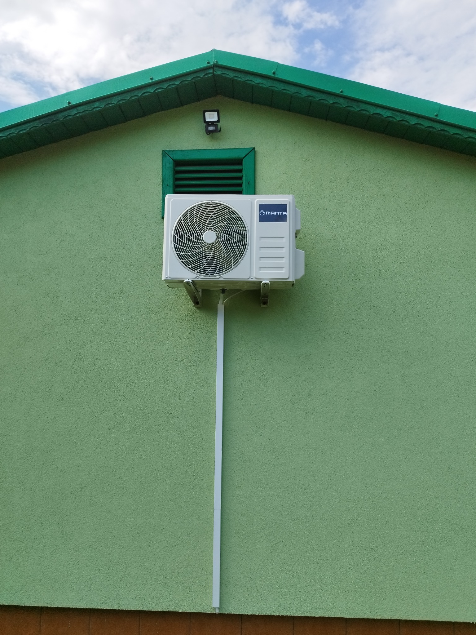 Biały klimatyzator Manta zamontowany na zielonej ścianie budynku z zielonym dachem i oknem. Widoczna biała rurka odprowadzająca skropliny.