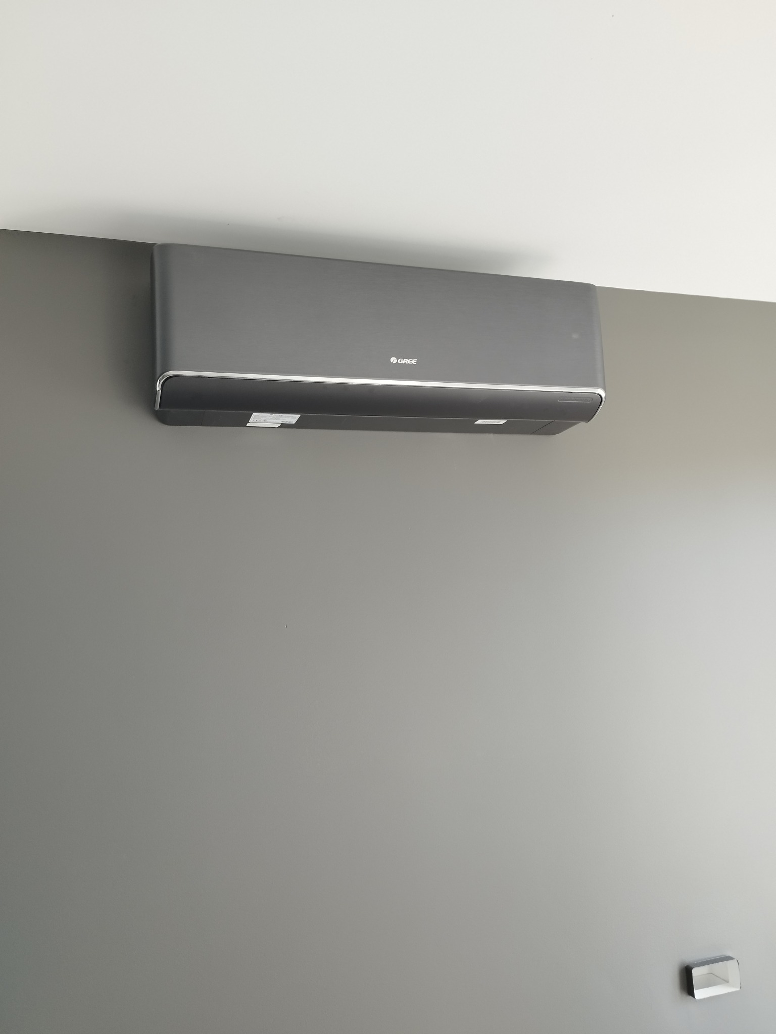 Szary klimatyzator marki Gree zamontowany na szarej ścianie w pomieszczeniu o minimalistycznym wystroju, z widocznym fragmentem sufitu.
