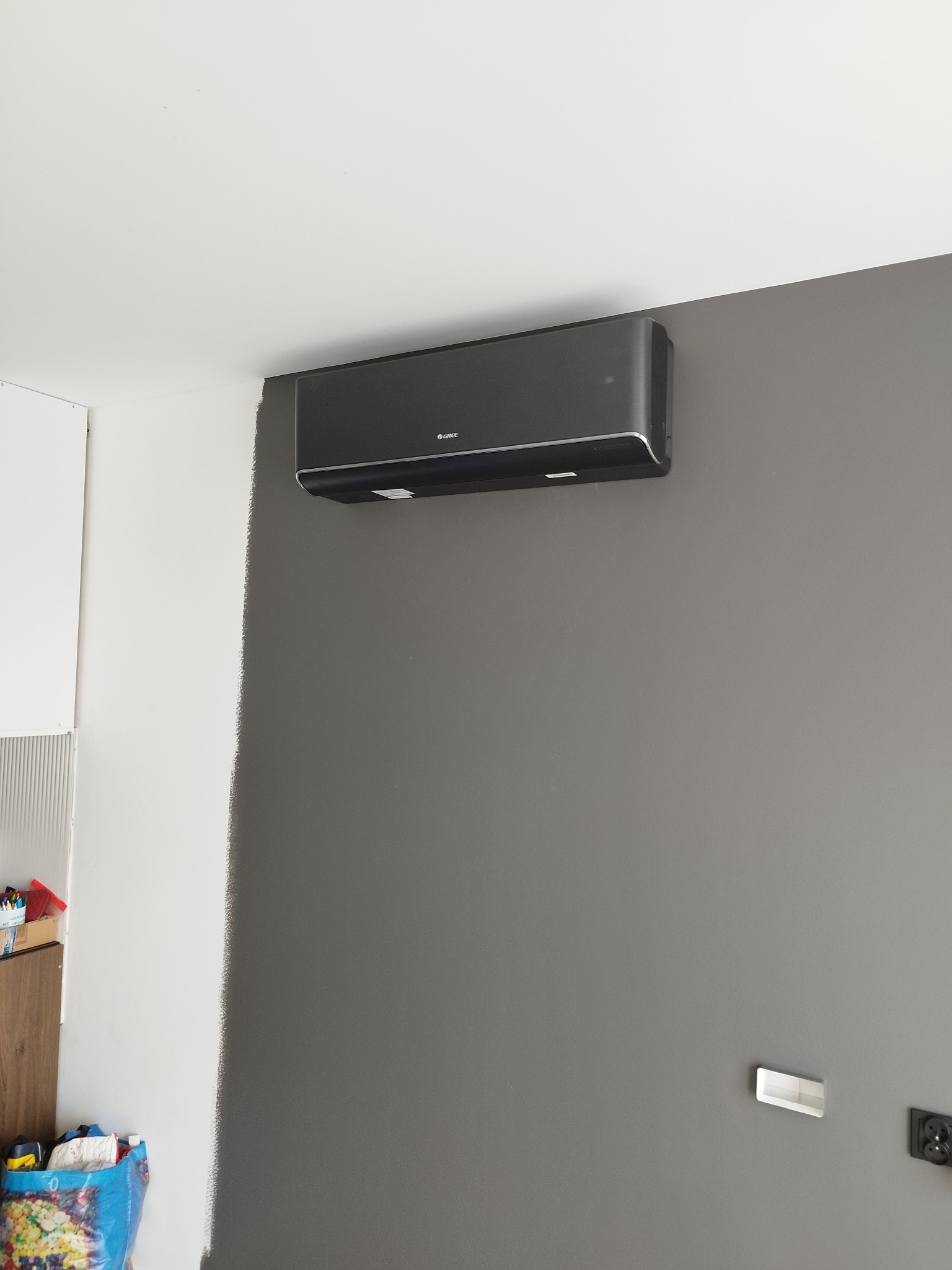 Czarny klimatyzator Gree zamontowany na szarej ścianie w pomieszczeniu z elementami białej ściany i widocznym fragmentem mebli. Widoczne gniazdka elektryczne.