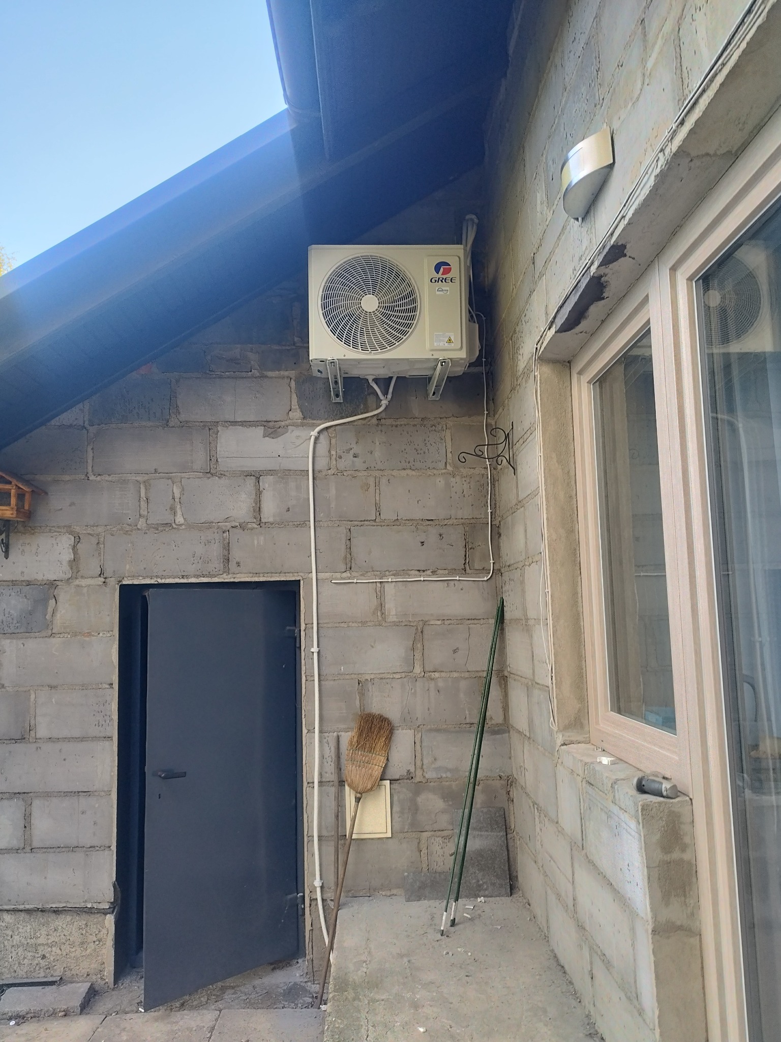Zamontowana klimatyzacja GREE na szarej ścianie z bloków obok okna i ciemnych drzwi. Na ścianie widoczna instalacja elektryczna i rurki doprowadzające czynnik chłodniczy.