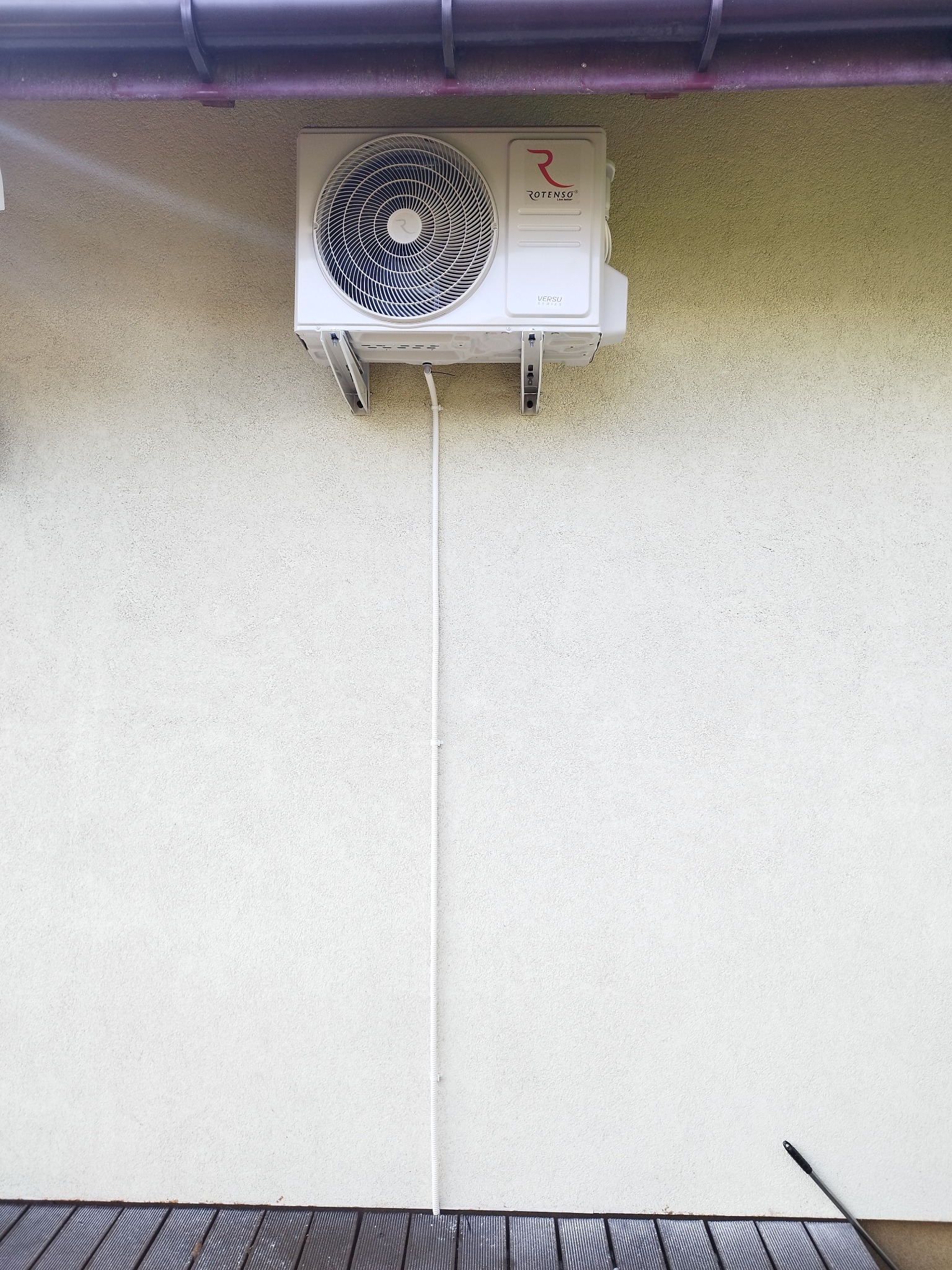 Biała jednostka zewnętrzna klimatyzacji Rotenso zamontowana na jasnej elewacji budynku z widoczną instalacją. Ujęcie z dołu, fragment dachu i desek podłogowych.