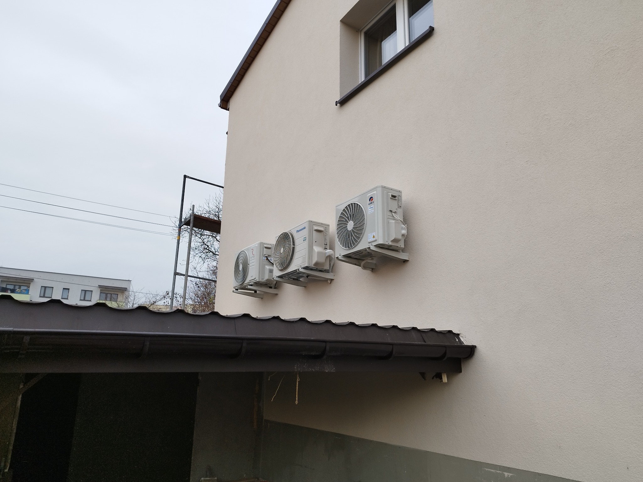 Trzy klimatyzatory różnych marek zamontowane na elewacji jasnego budynku, widoczne rynny i fragment rusztowania. Ujęcie z dołu, pochmurne niebo.