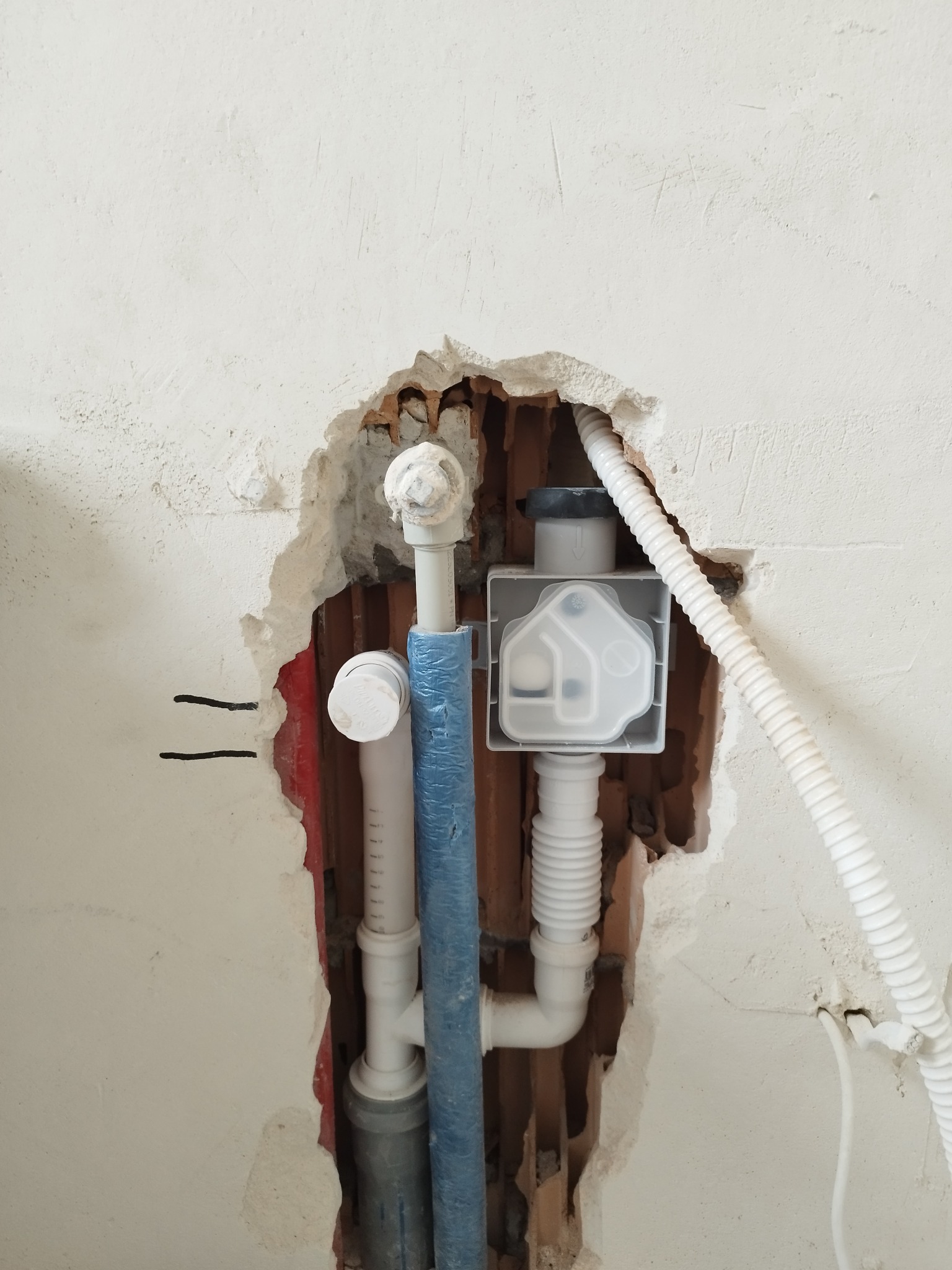 Instalacja wodno-kanalizacyjna w ścianie podczas montażu. Widoczne rury, kształtki i biały przewód elektryczny w korytarzu. Fragment ściany jest uszkodzony.