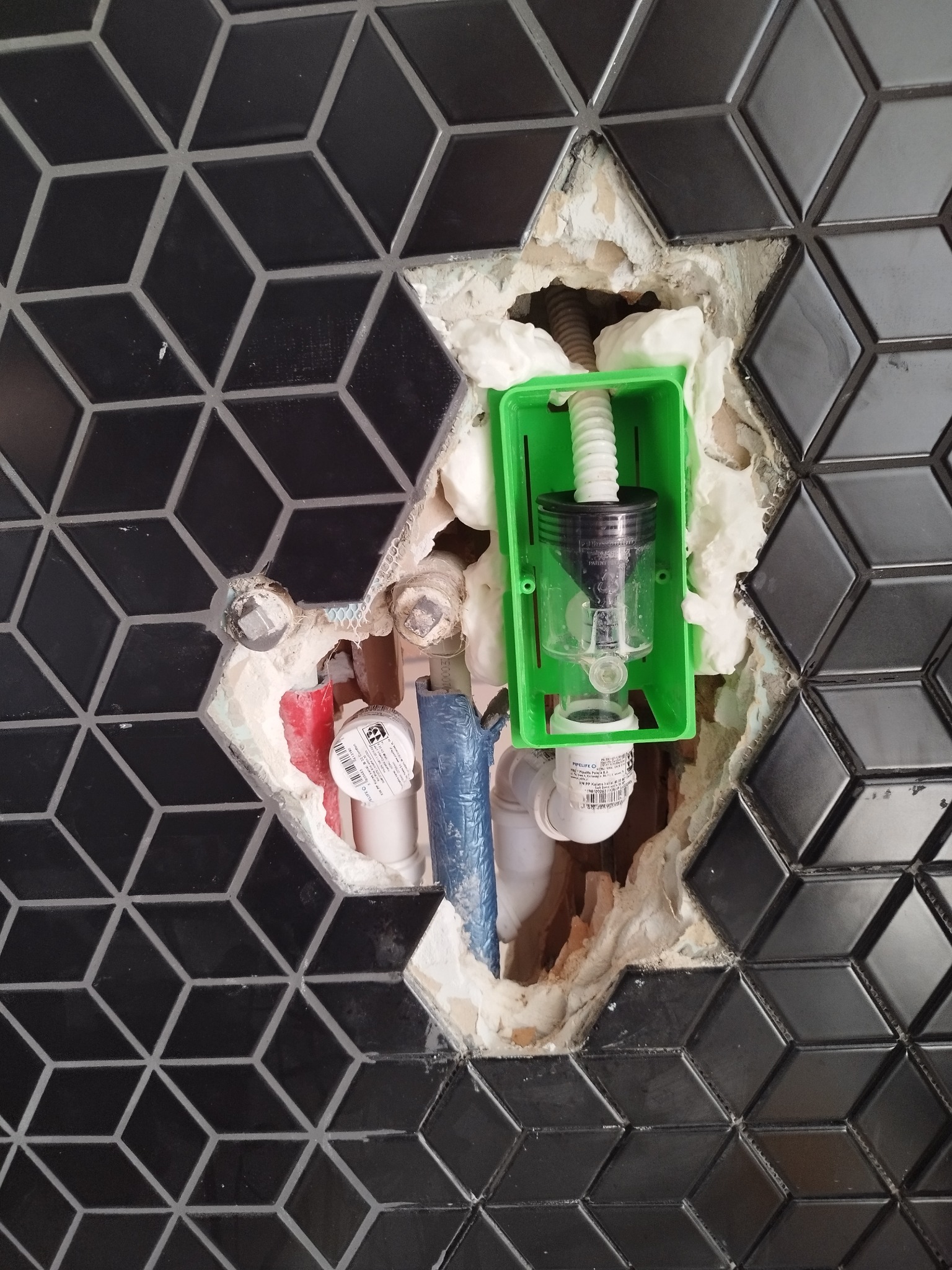 Fragment instalacji hydraulicznej w ścianie z czarną, geometryczną mozaiką, zabezpieczonej pianką montażową i zieloną obudową. Widoczne rury i elementy mocujące.