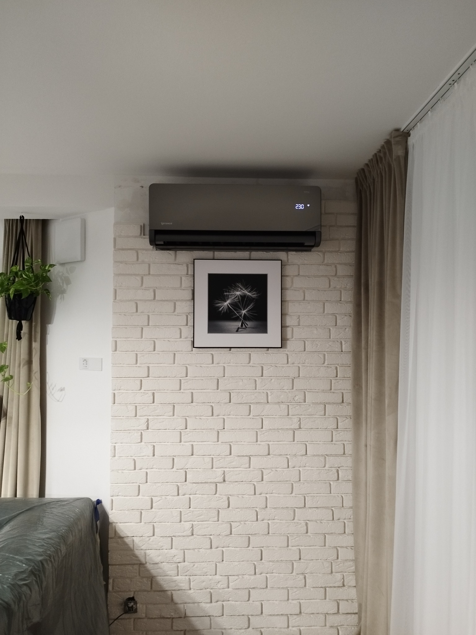Szary klimatyzator Rotenso zamontowany na ścianie z białej cegły, pod nim czarno-biała grafika w ramce. Widoczne zasłony i roślina doniczkowa.