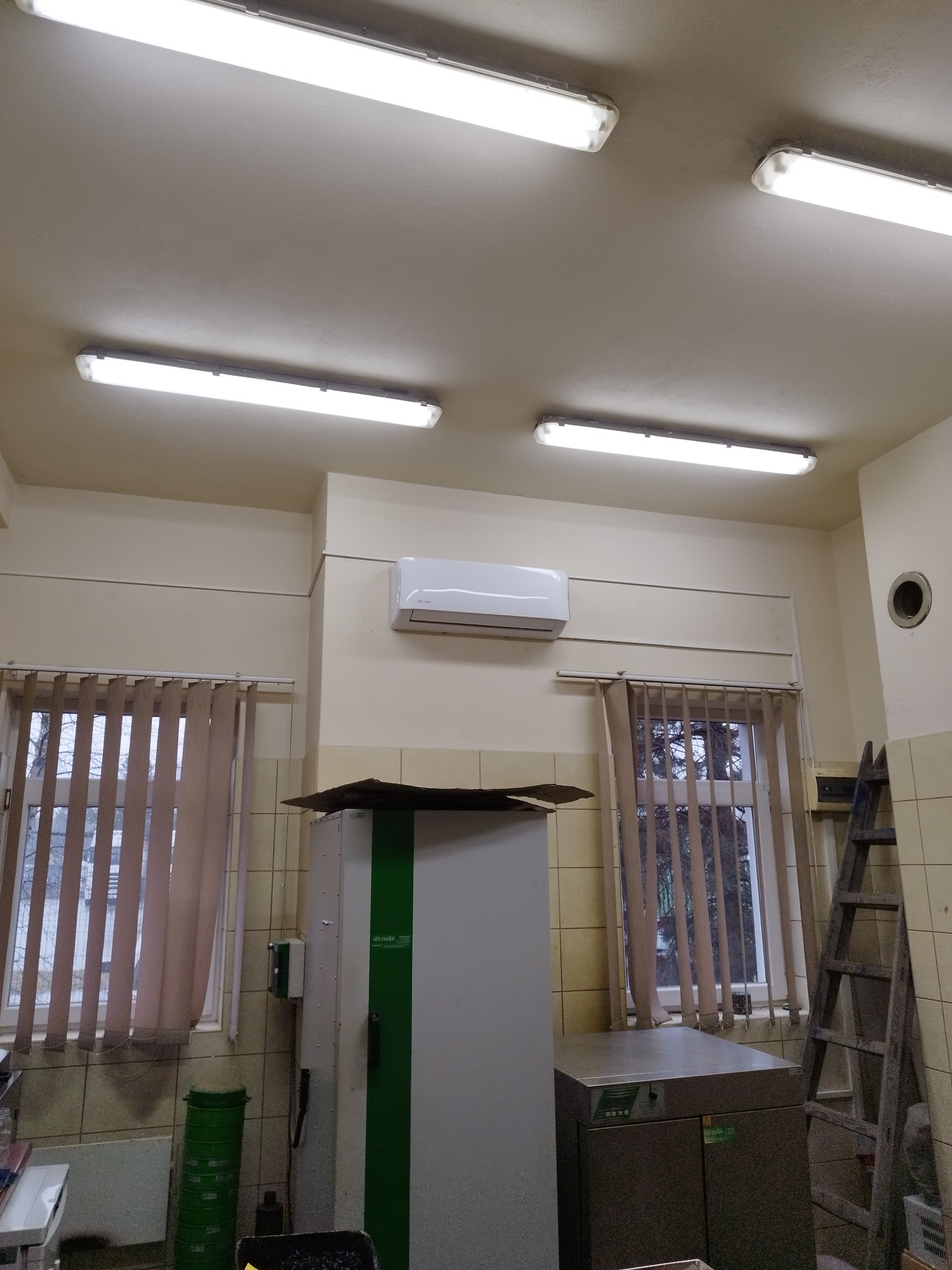 Biała klimatyzacja ścienna zamontowana w pomieszczeniu z oknami w tle. Widoczne lampy sufitowe, drabina i inne urządzenia.