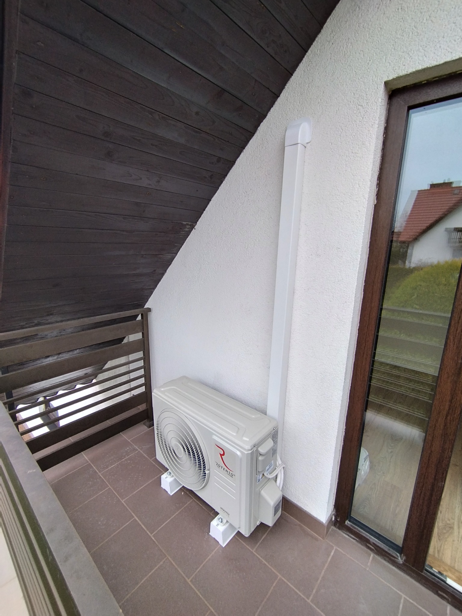 Biała klimatyzacja Rotenso na balkonie z brązową podłogą, zamontowana na białych wspornikach, z białą rurą odprowadzającą. Widok z góry na dom z czerwonym dachem.