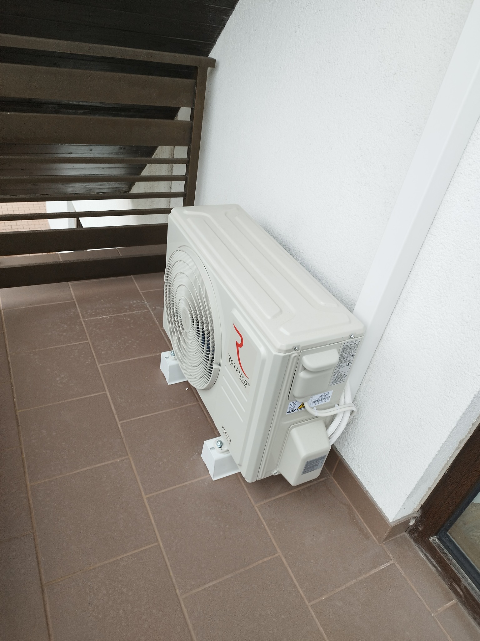 Kompaktowa jednostka zewnętrzna klimatyzacji Rotenso umieszczona na balkonie, z widocznymi mocowaniami i przewodami. Ściana o fakturze baranka i brązowe płytki podłogowe.