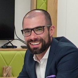 Marcin Cholewa Freedom Nieruchomości - Uśmiechnięty mężczyzna w okularach i granatowej marynarce z fioletową poszetką, siedzący w biurze z zielonymi akcentami, prezentujący się profesjonalnie.