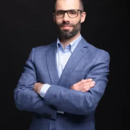 Portret mężczyzny w okularach i niebieskiej marynarce, z założonymi rękami na ciemnym tle. Profesjonalny wizerunek agenta nieruchomości.