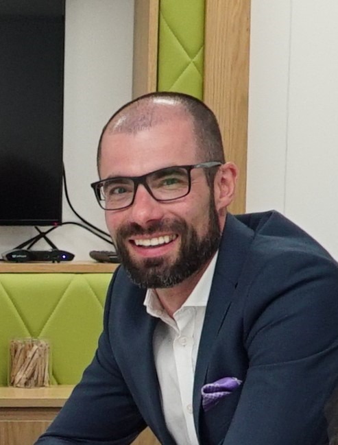 Uśmiechnięty mężczyzna w okularach i granatowej marynarce z fioletową poszetką, siedzący w biurze z zielonymi akcentami, prezentujący się profesjonalnie.