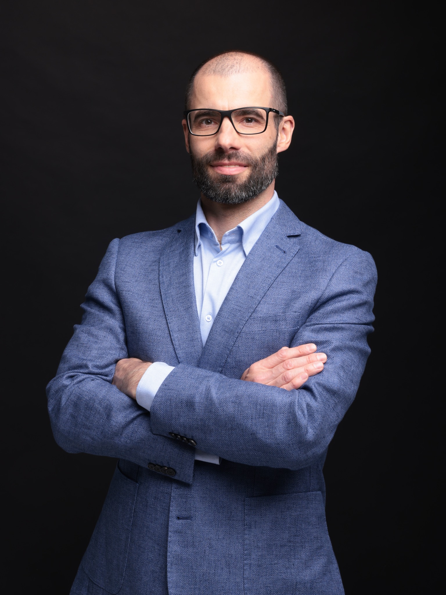 Portret mężczyzny w okularach i niebieskiej marynarce, z założonymi rękami na ciemnym tle. Profesjonalny wizerunek agenta nieruchomości.