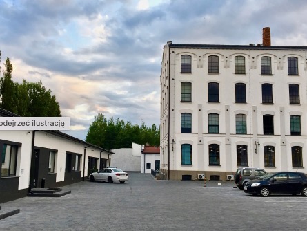 Widok na odnowiony budynek fabryczny z ceglanym kominem, z parkingiem wyłożonym kostką brukową, na którym stoją samochody osobowe.