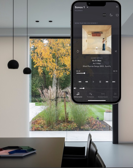 Nowoczesne wnętrze z widokiem na jesienny ogród, w kadrze smartfon z aplikacją Sonos odtwarzającą utwór 'As It Was' Harry'ego Stylesa, widoczne wiszące lampy kuliste i minimalistyczny stół...